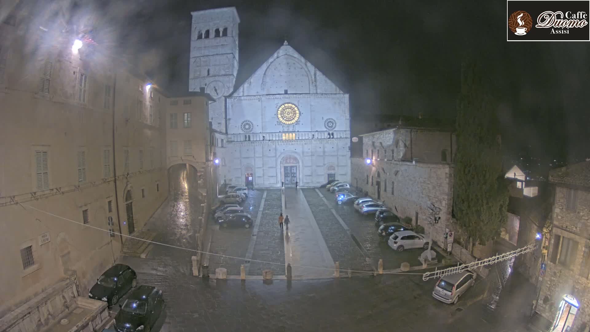 A large stone cathedral stands in a town square at night during a rainstorm, surrounded by parked cars and older buildings.