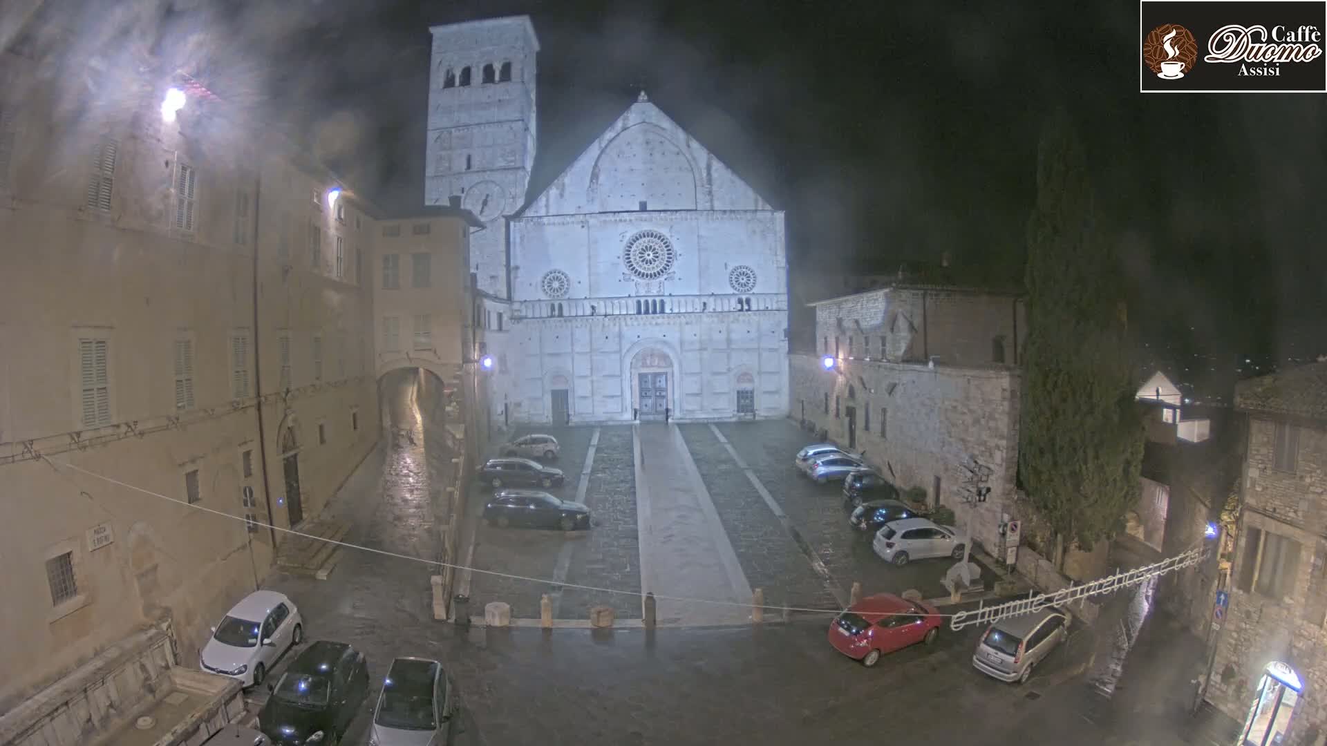 A large, light-colored cathedral stands in a town square at night, under a misty rain, surrounded by parked cars and stone buildings.