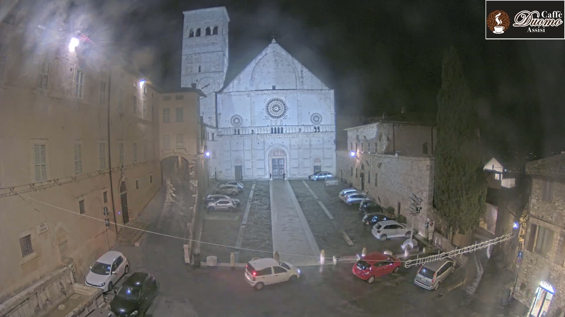 A nighttime view of a town square with a large cathedral as its centerpiece, cars parked around the square, and light rain falling.