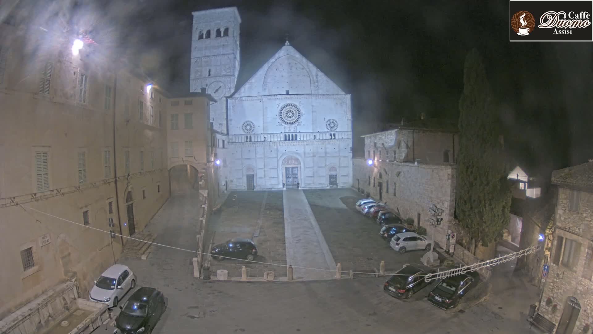 A nighttime view of a large cathedral in a town square with several cars parked, under a cloudy sky.