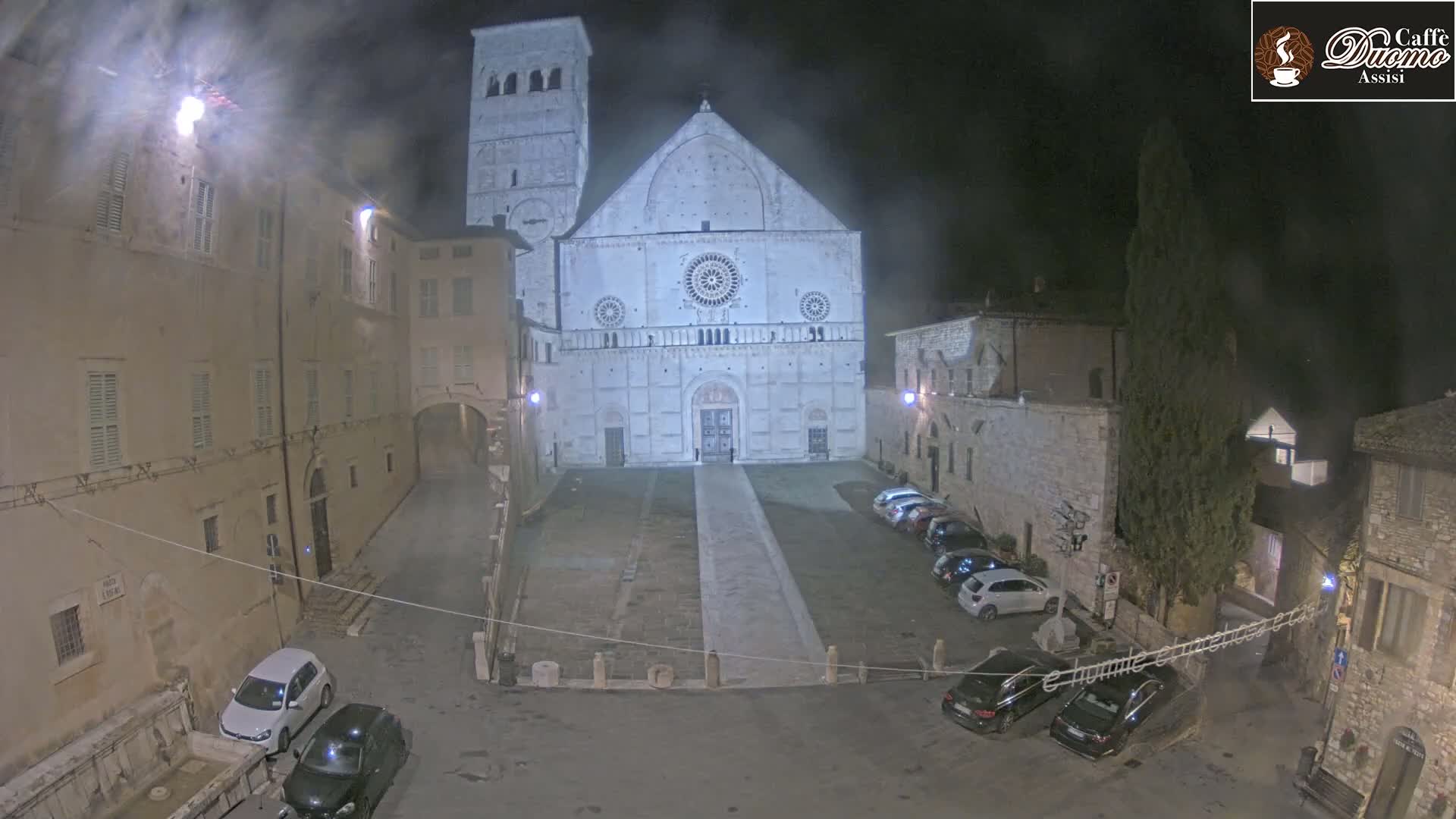 A large, light-colored cathedral stands in a dimly lit, empty town square at night, surrounded by older stone buildings and a few parked cars.