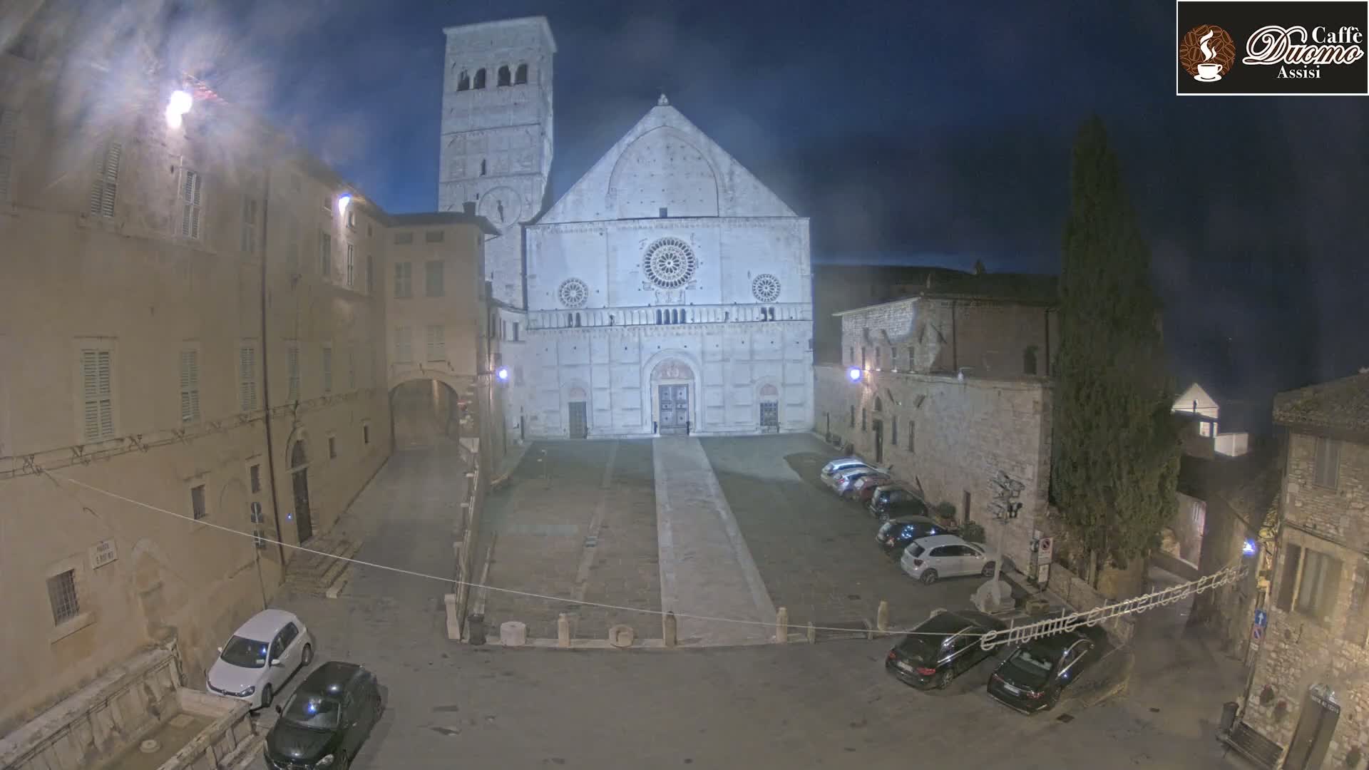 A large stone cathedral is illuminated at night in a town square where several cars are parked, under a dark, clear sky.