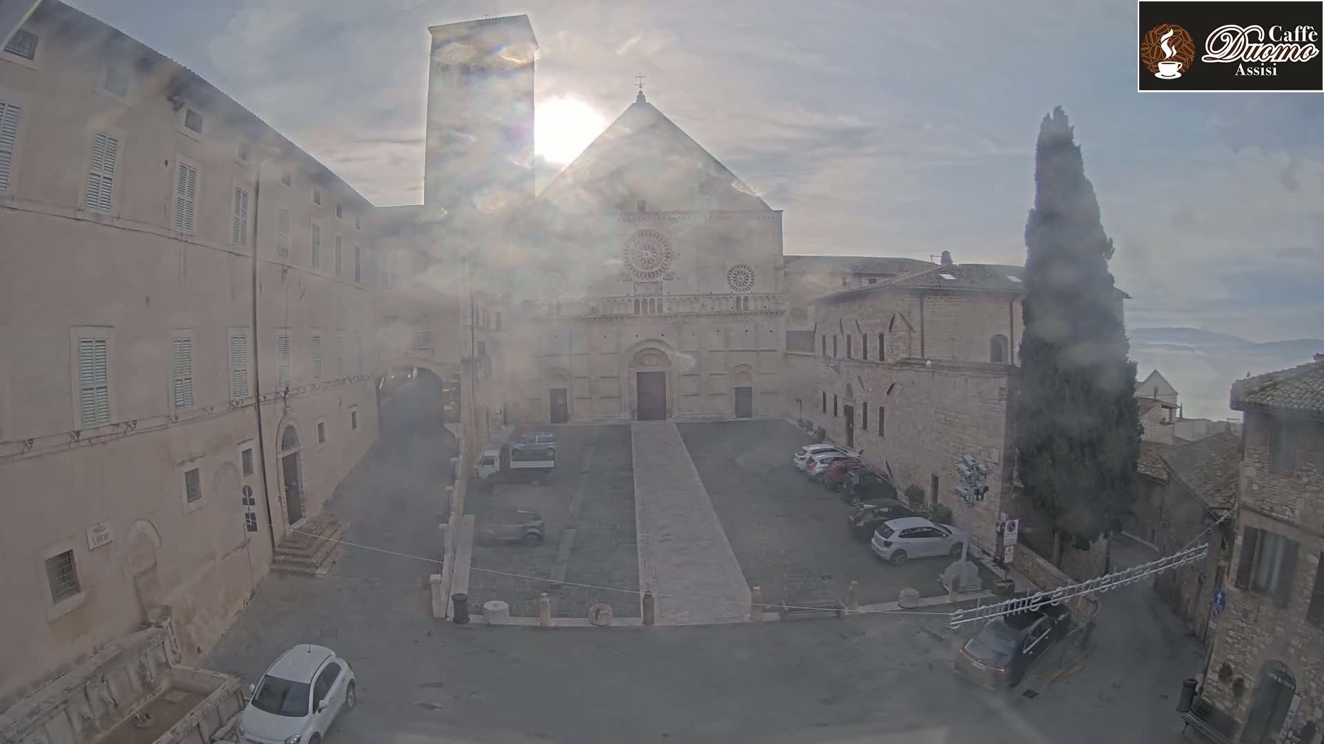 A hazy sun shines over a stone plaza and cathedral, with cars parked in the square and surrounding buildings visible under a mostly cloudy sky.