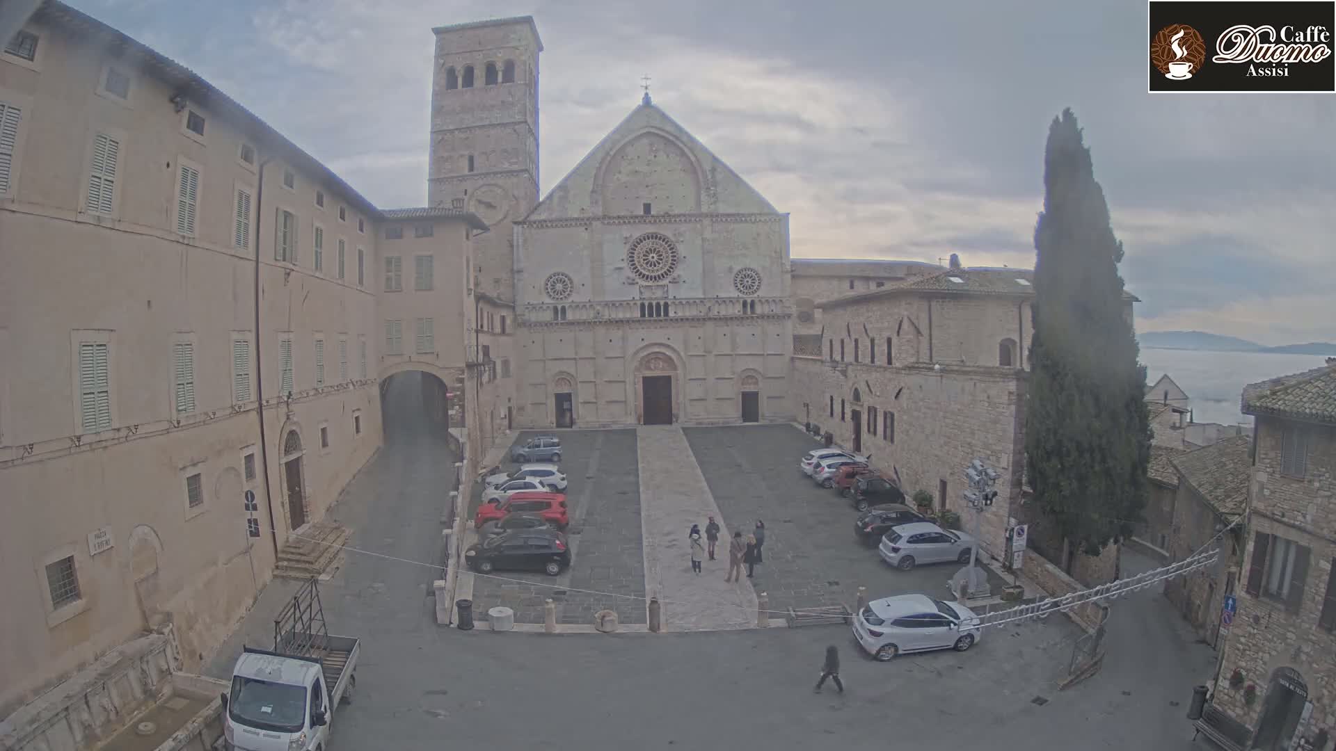 A large stone cathedral faces a plaza with several cars parked, surrounded by older buildings under an overcast sky.