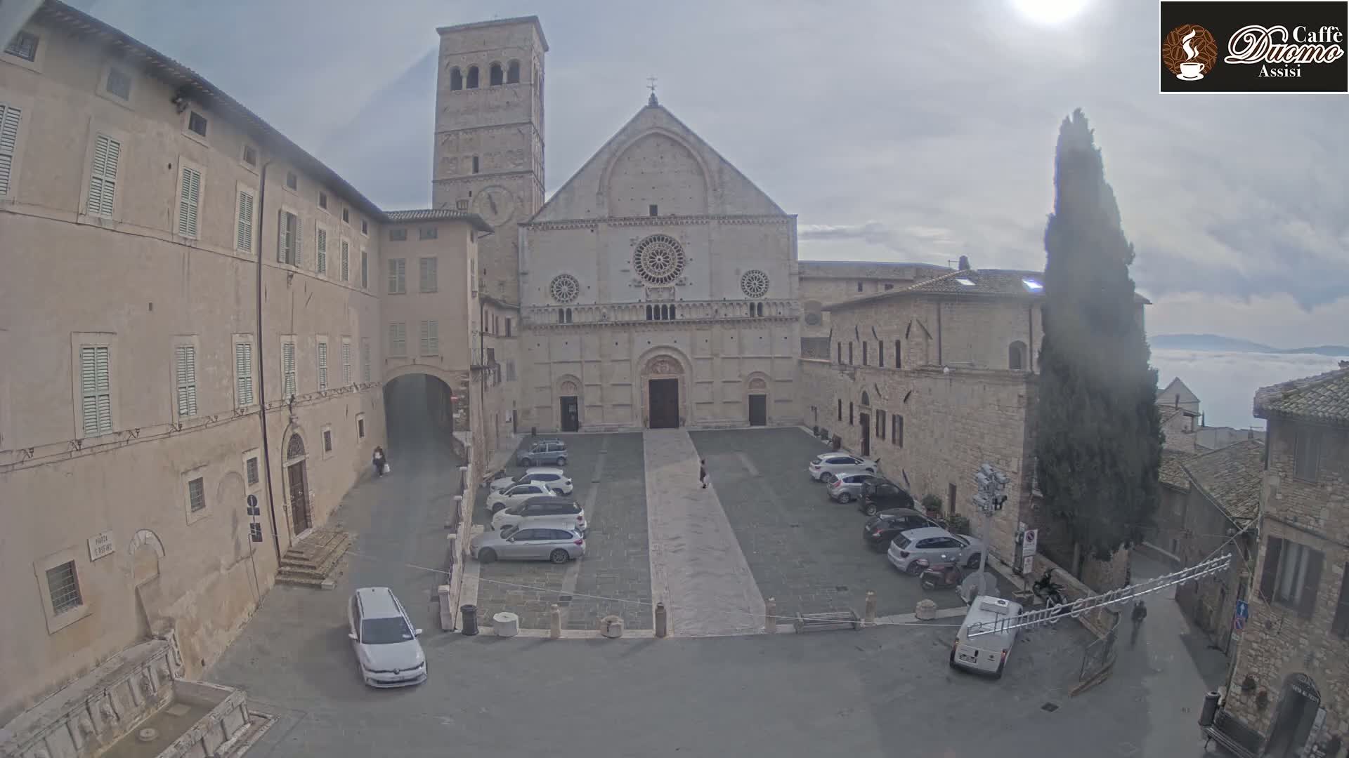 A large stone church and surrounding buildings are visible in a city square with several cars parked, under a mostly cloudy sky with a distant layer of fog.