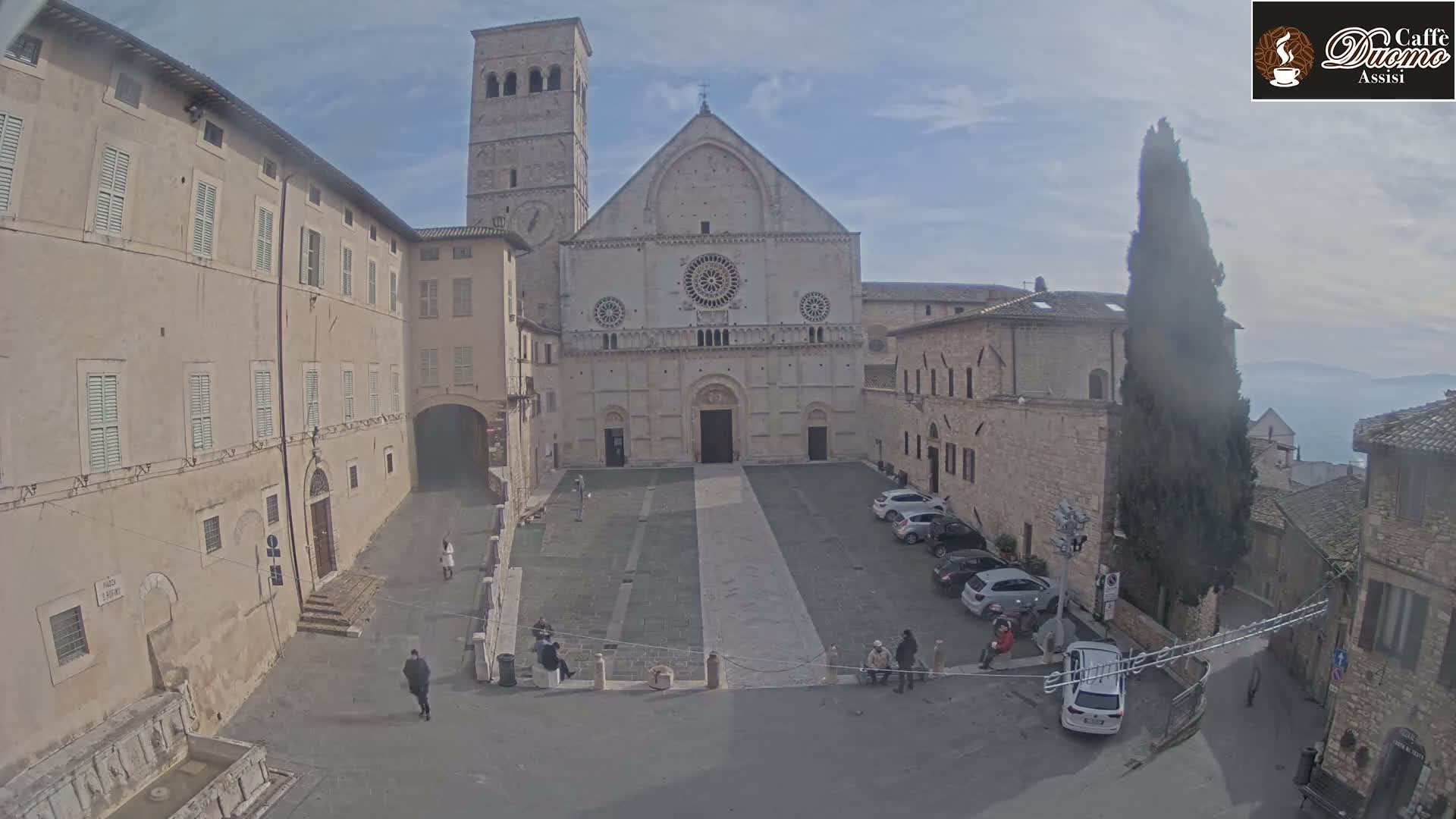 A large stone church stands in a town square on an overcast day, with several cars parked nearby and a few people walking around.
