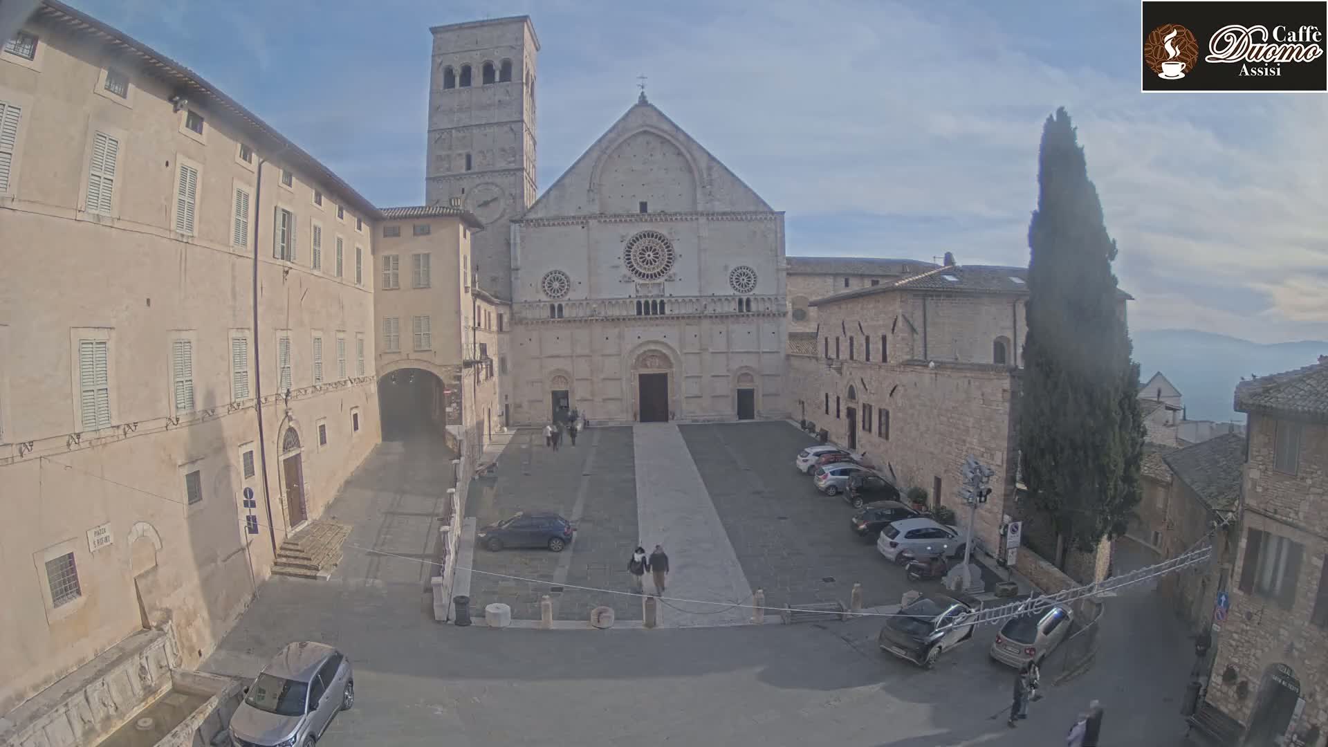 A large stone cathedral stands in a plaza surrounded by aged buildings under a mostly clear sky, with several cars parked nearby and a few pedestrians walking.