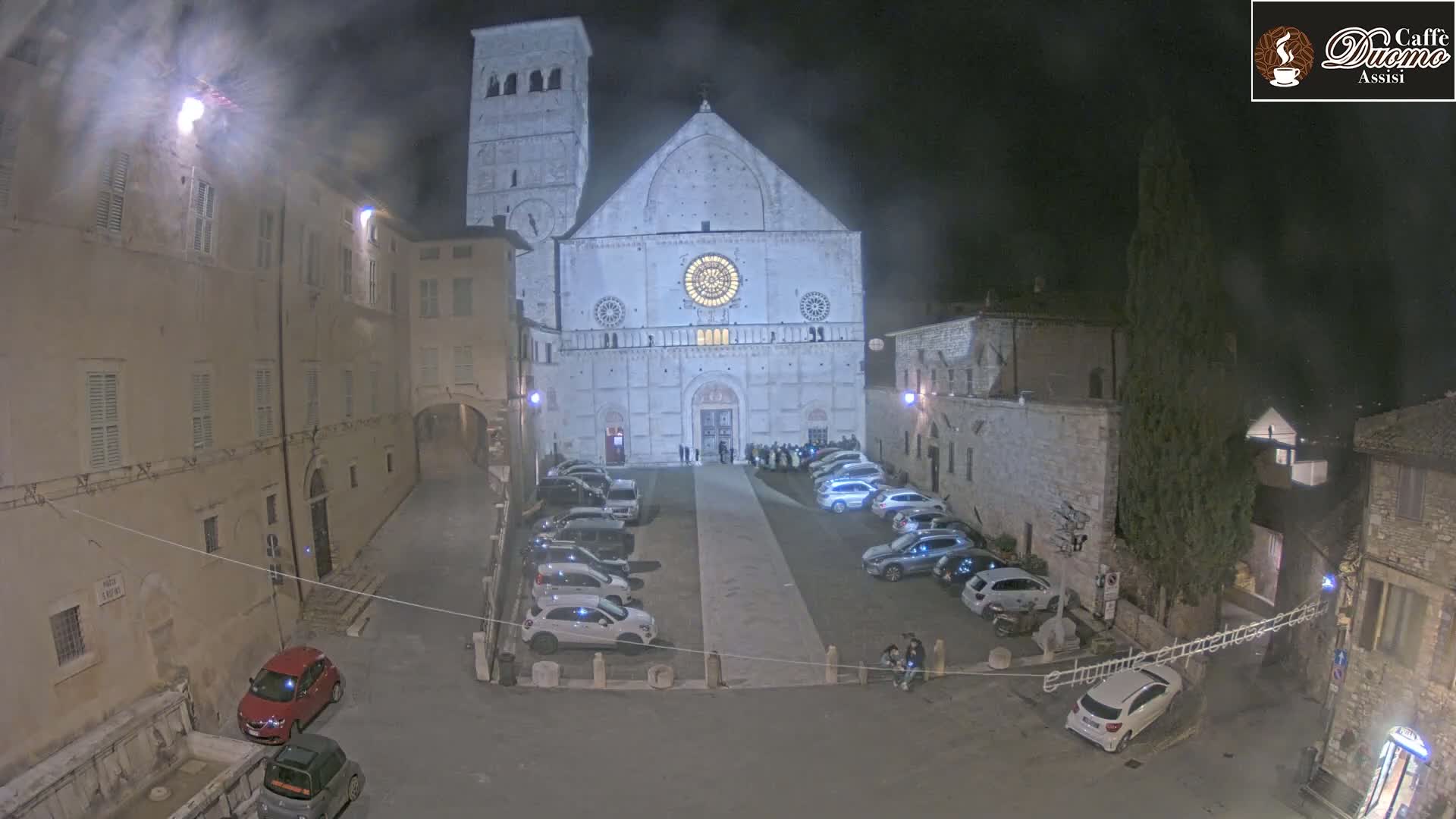 A nighttime view of a cathedral's facade and surrounding square, illuminated, with several cars parked, under hazy conditions.