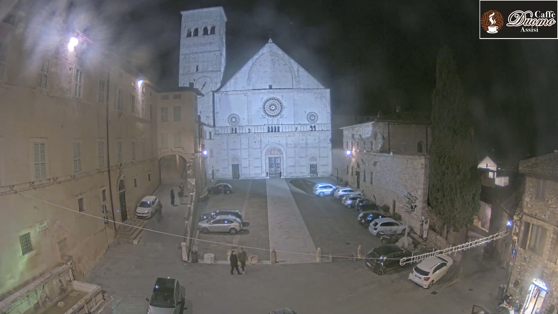 A large stone cathedral is seen at night in a town square with several cars parked, and a few pedestrians are walking nearby.