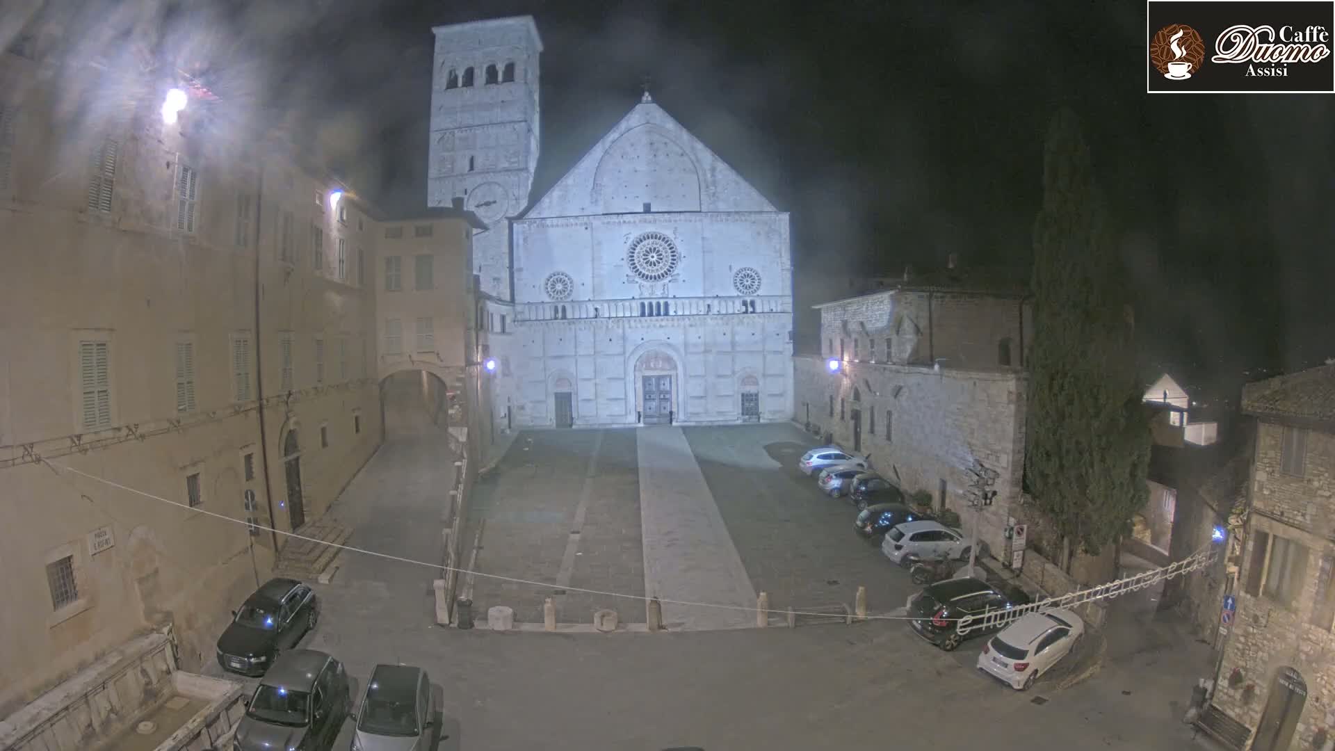 A nighttime, hazy view of a cathedral's facade and square, surrounded by stone buildings and parked cars.