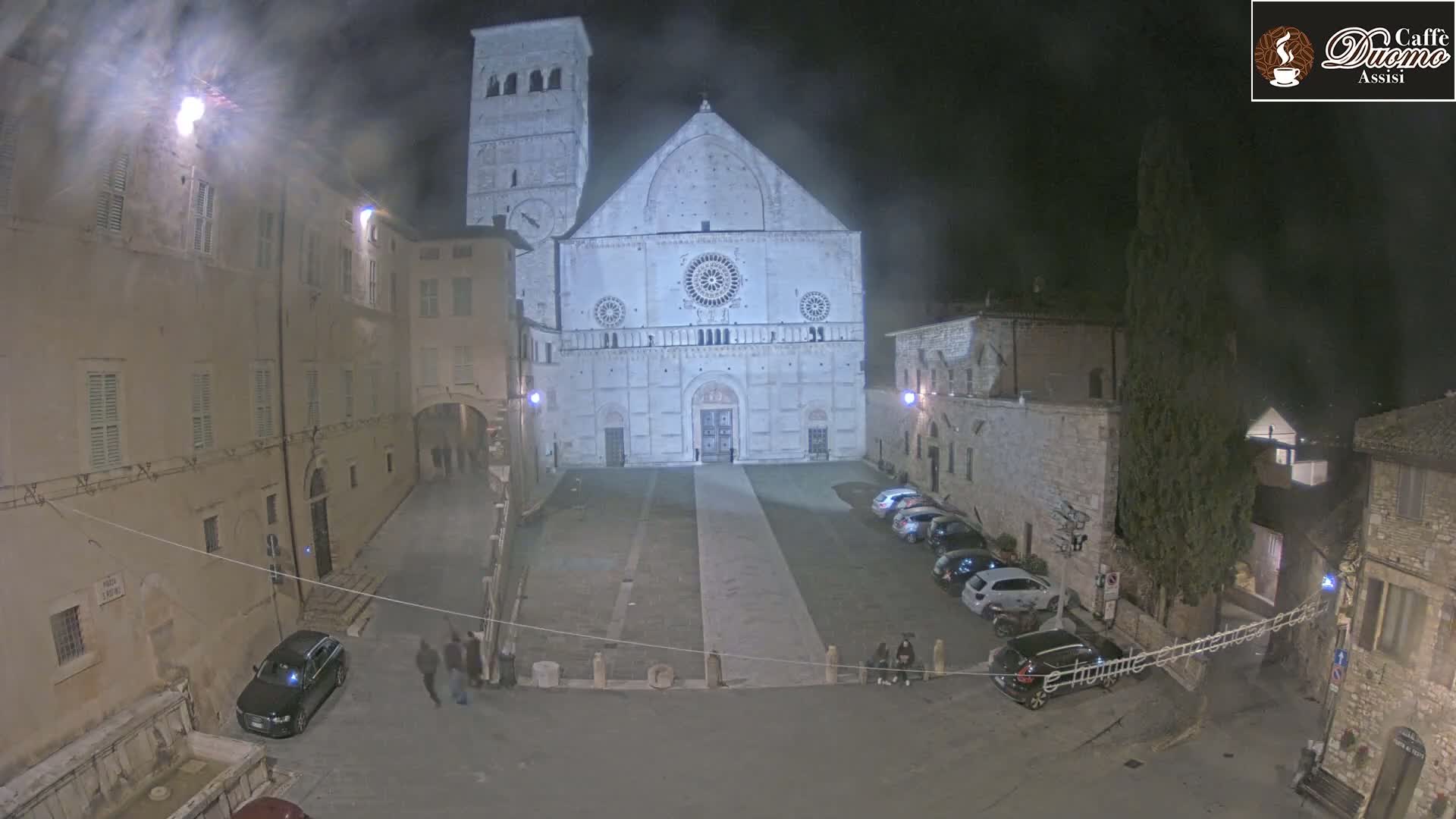 A nighttime, hazy view of a large stone cathedral in a town square with several cars parked and a few people walking around.