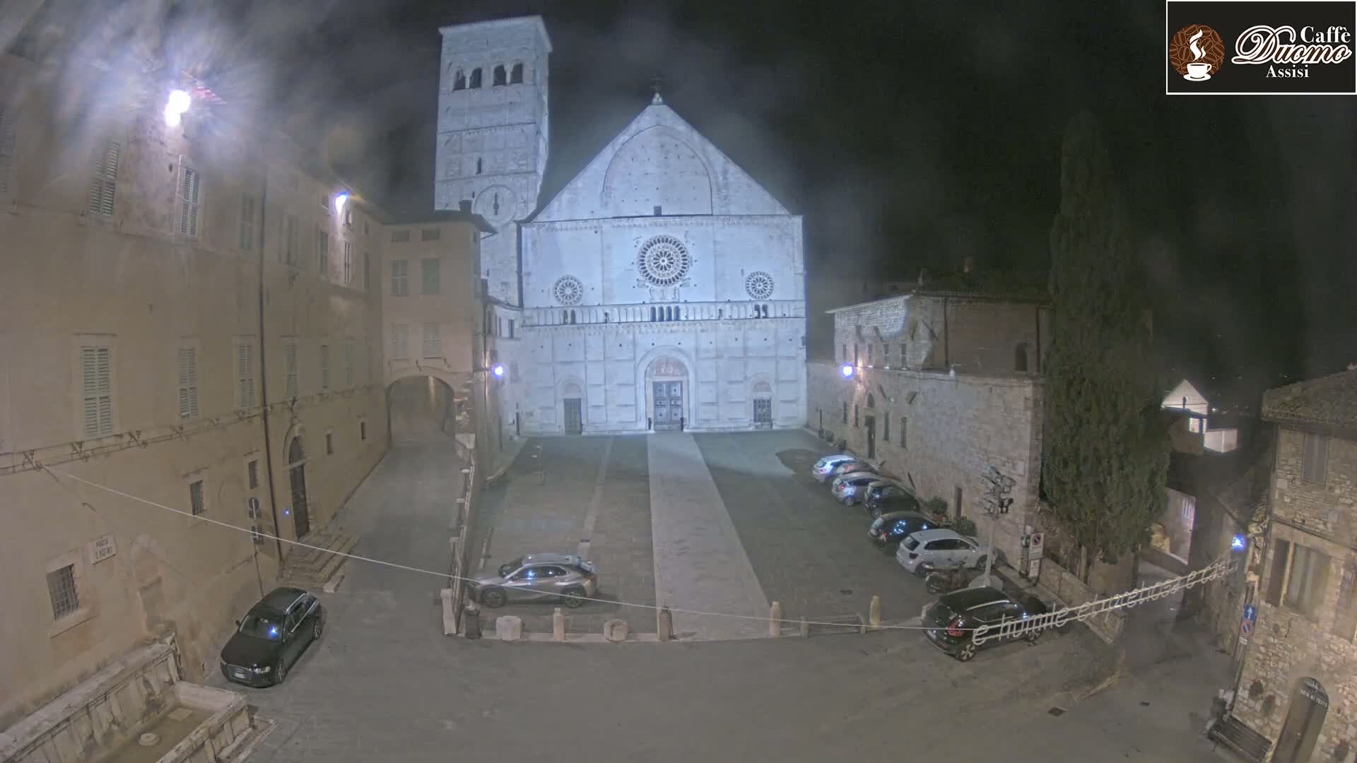 A large, light-colored cathedral is situated in a dimly lit, stone-walled plaza at night, with several cars parked in the square.