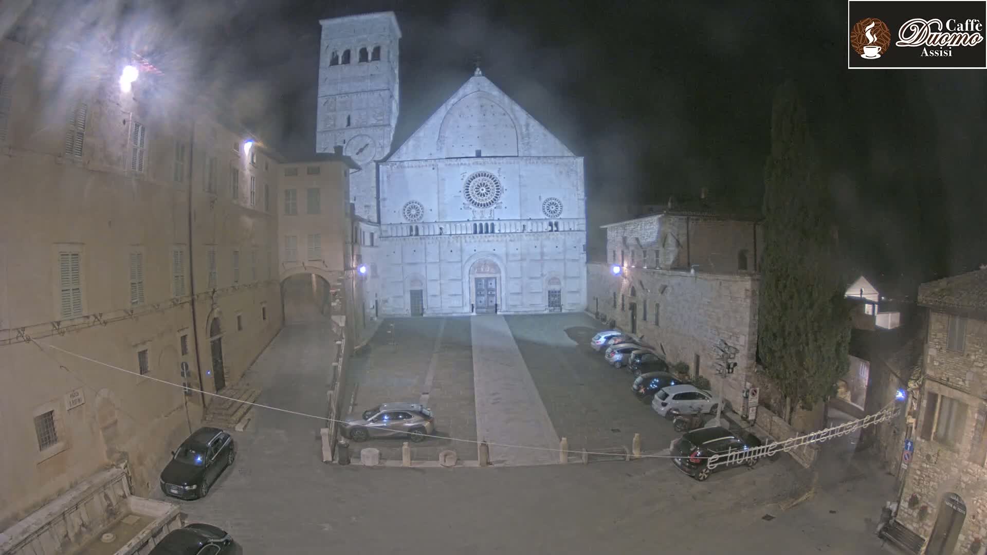 A large, light-colored cathedral stands in a dimly lit town square at night, surrounded by older stone buildings and several parked cars under hazy or foggy conditions.