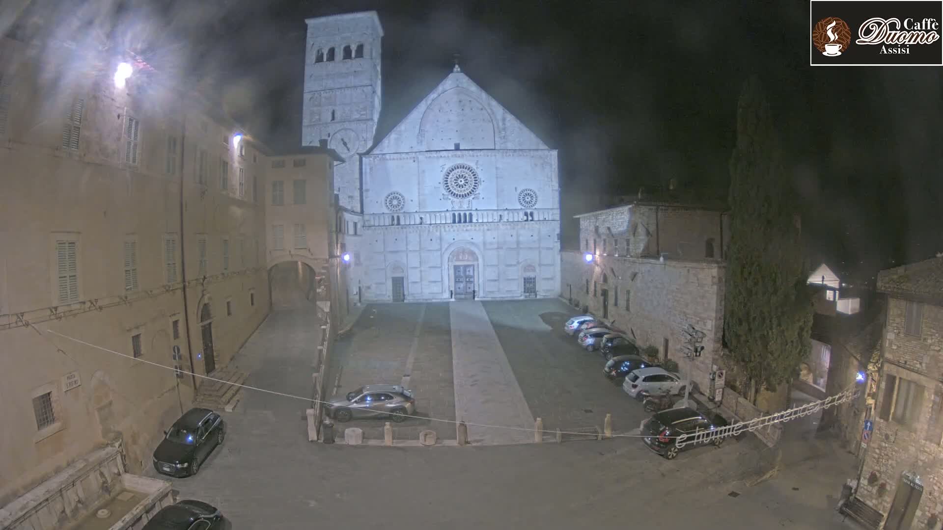 A large stone cathedral is visible at night in a town square with several cars parked, under a slightly hazy sky.