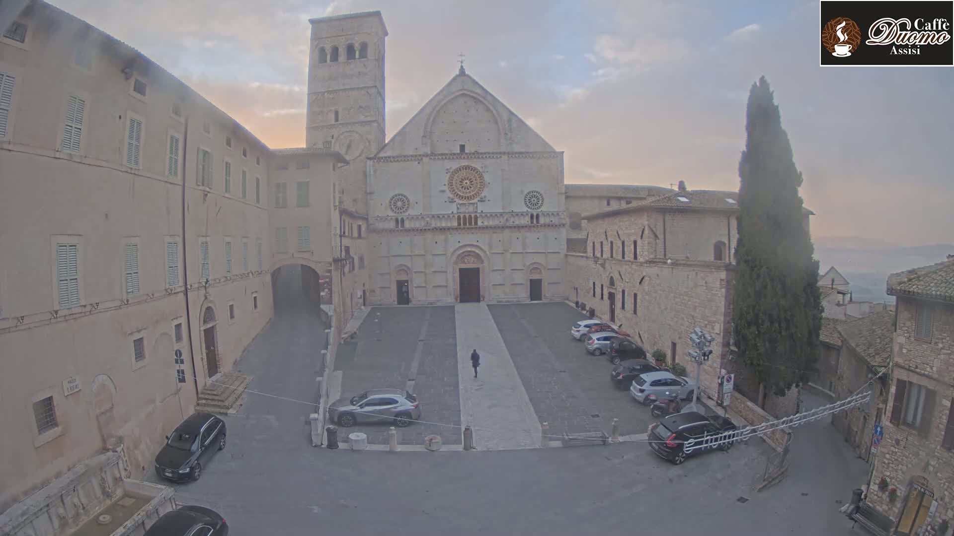 A person walks across a stone plaza in front of a large cathedral, surrounded by old buildings under a hazy sky.