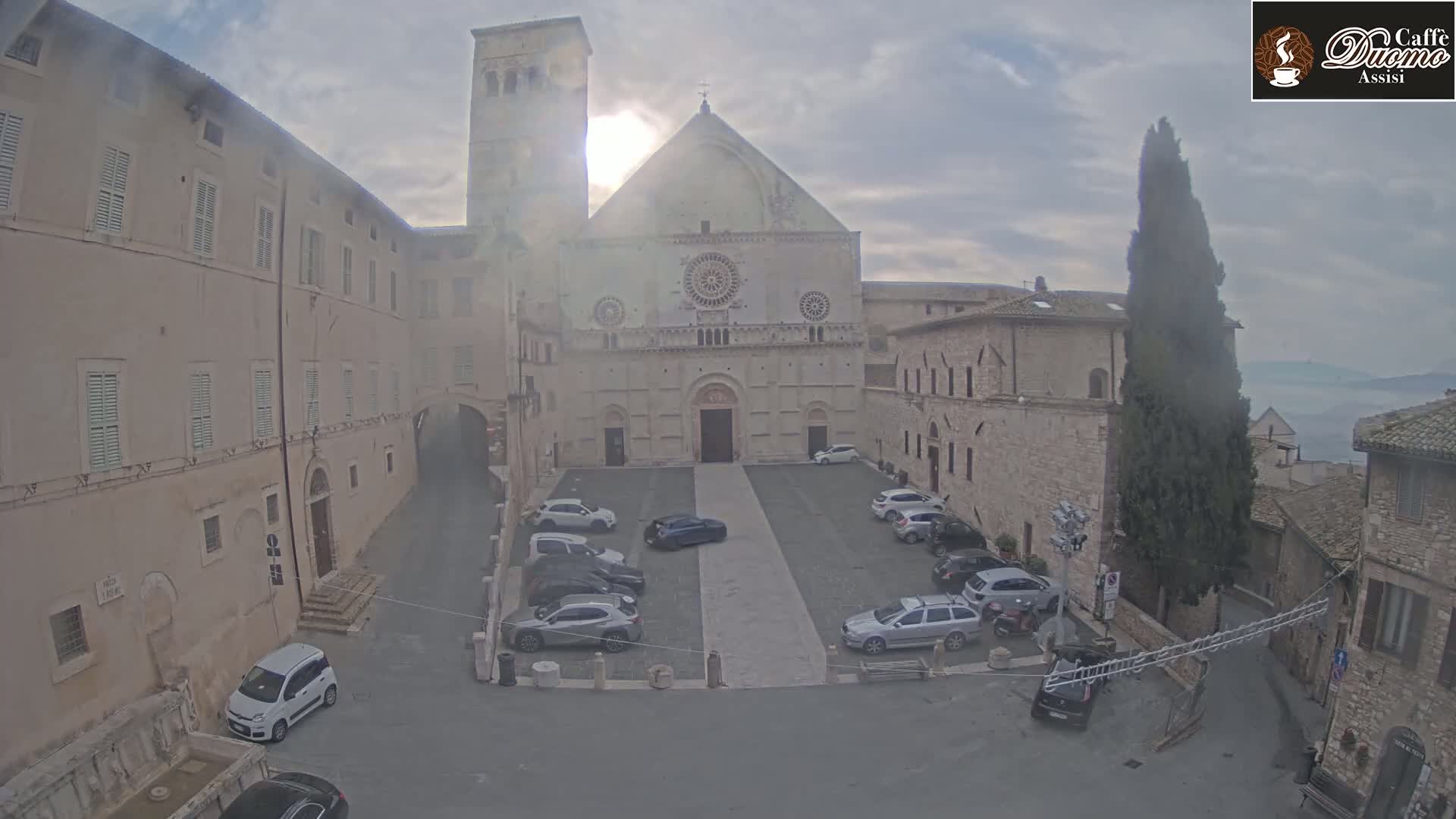 A stone cathedral and surrounding buildings, with cars parked in a square, under an overcast sky.