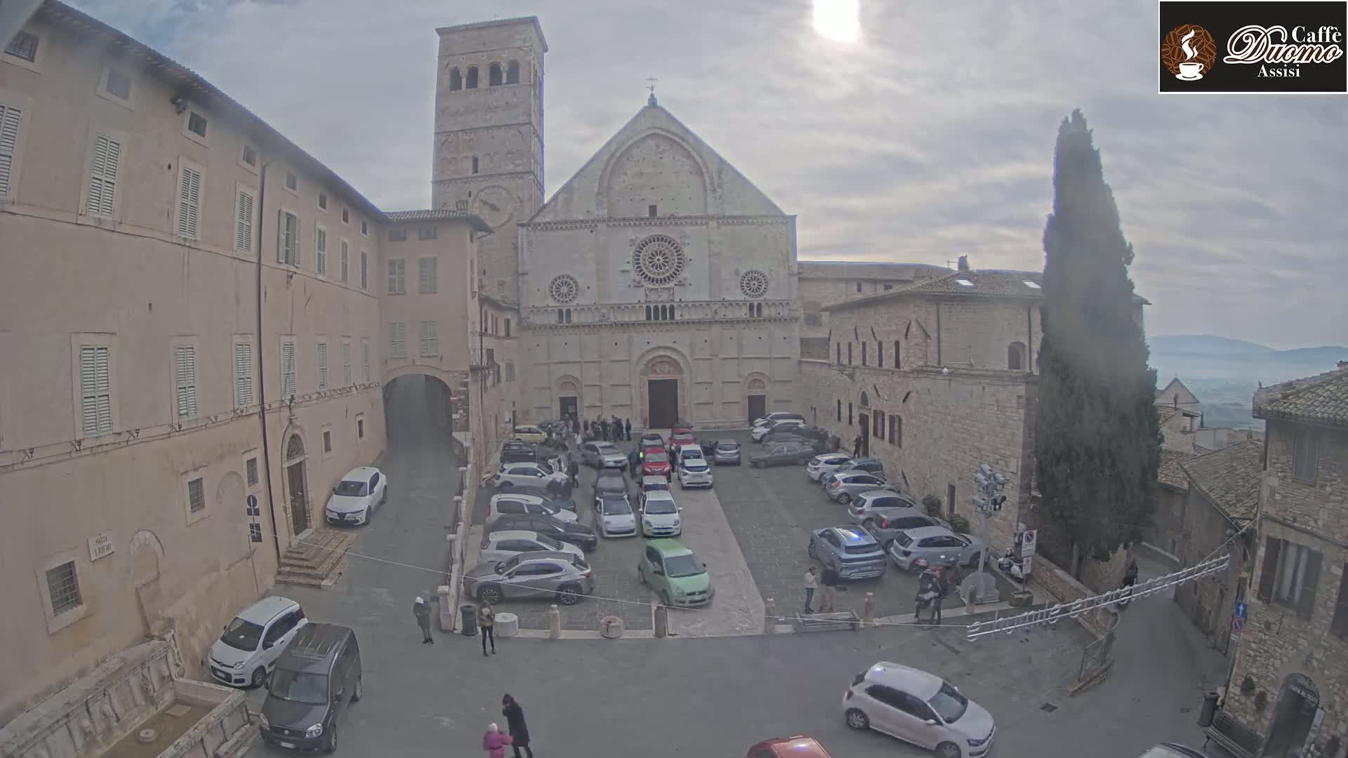 A large stone cathedral sits in a town square filled with parked cars under an overcast sky.