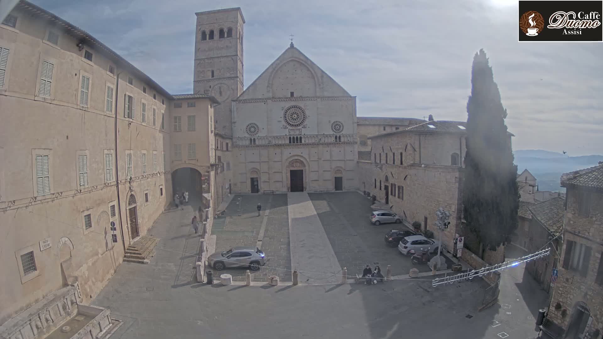 A large stone cathedral sits in a town square on a mostly sunny day, surrounded by other stone buildings and a few cars parked nearby.