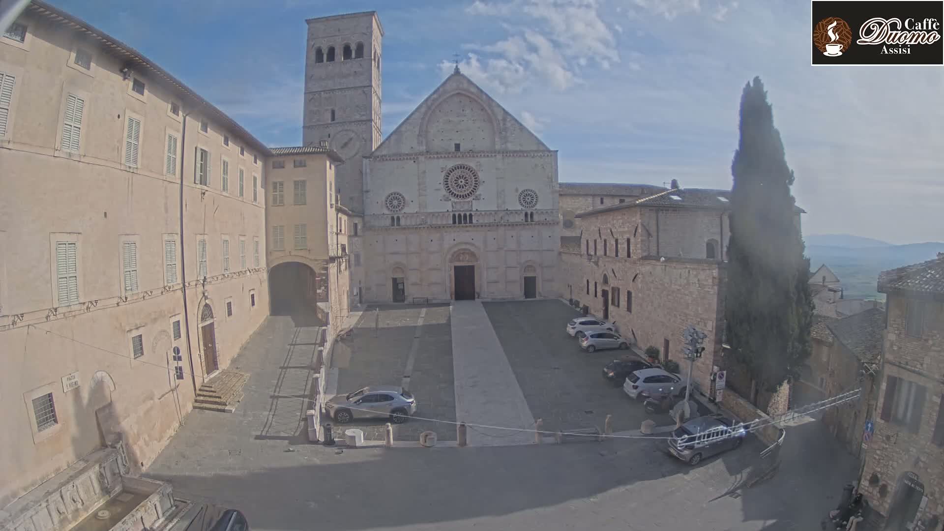 A large stone cathedral is situated in a town square, with several cars parked nearby, under a mostly clear sky.