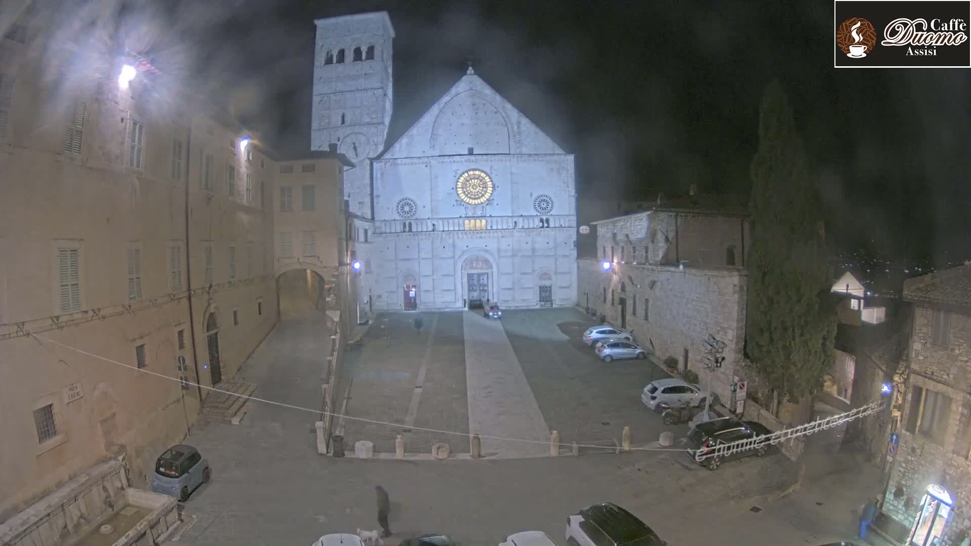 A large stone cathedral stands in a dimly lit town square at night, under a hazy sky.