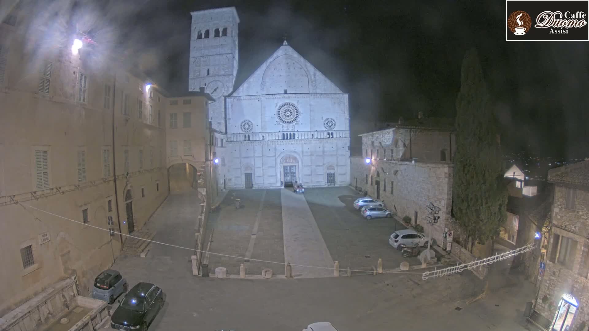A large stone cathedral is illuminated at night in a square surrounded by buildings, under hazy conditions.