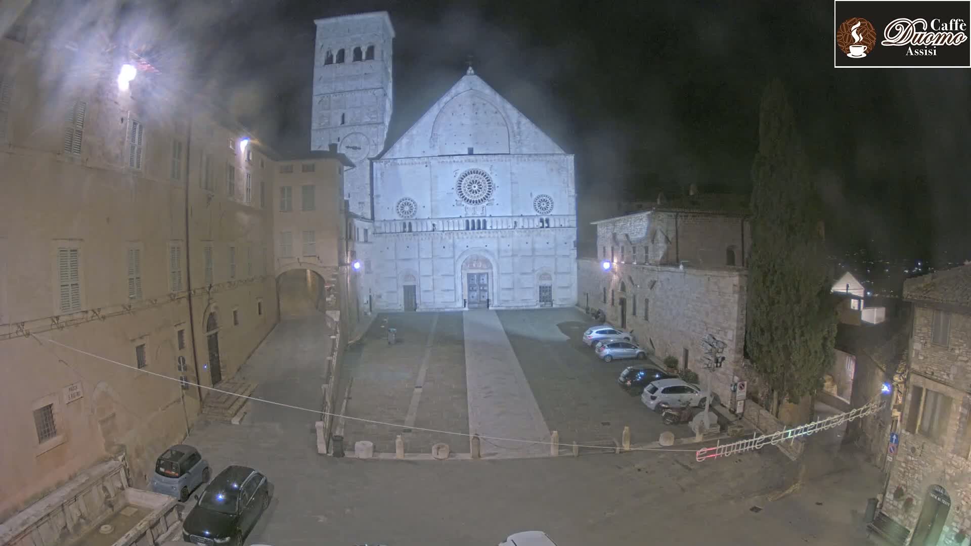 A large stone cathedral is prominently displayed in a town square at night, under hazy conditions, with several cars parked nearby and older buildings surrounding the square.