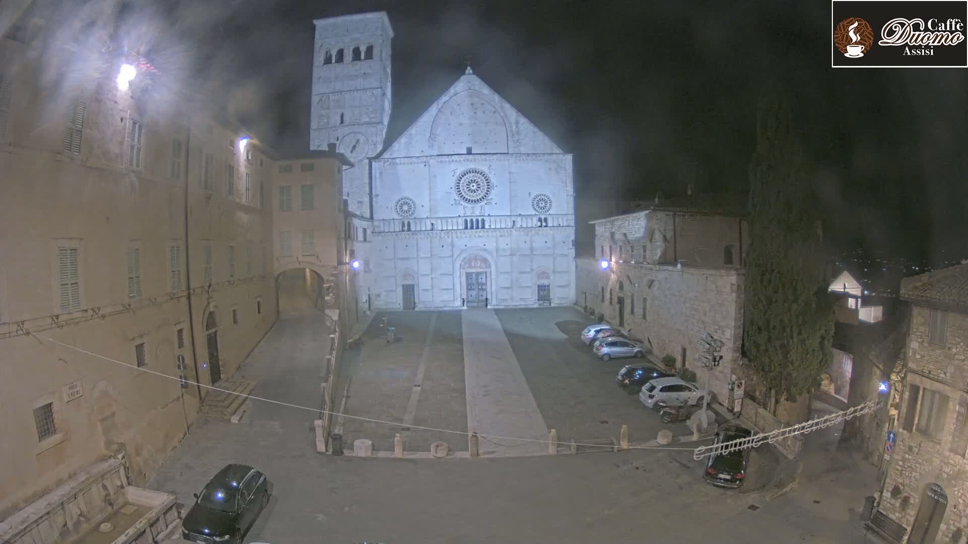 A nighttime view of a large stone cathedral in a town square, with several cars parked and a few buildings surrounding the square under hazy conditions.