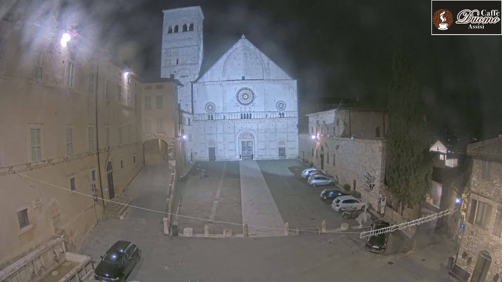 A large, light-colored cathedral stands in a dimly lit, empty plaza at night, surrounded by older stone buildings under hazy conditions.