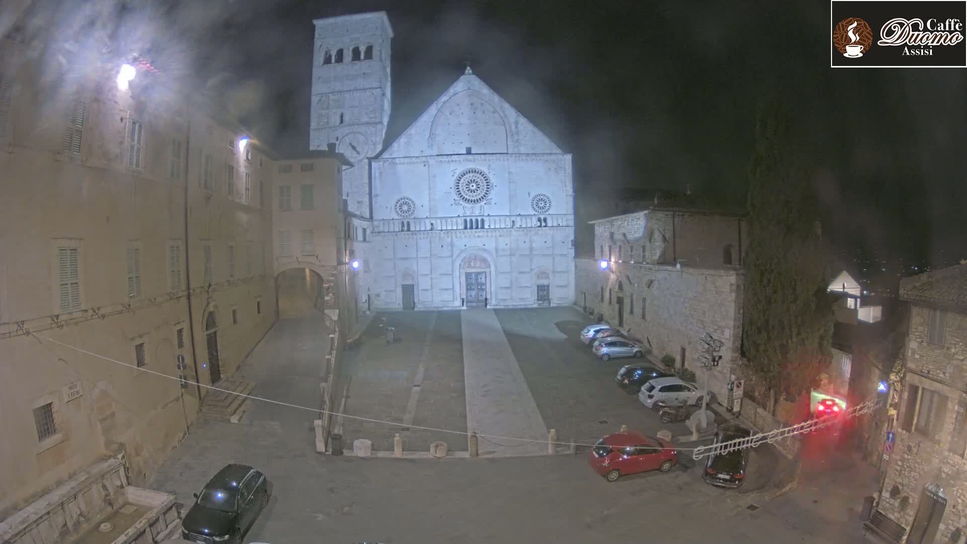 A nighttime view of a large, light-colored cathedral in a town square with several cars parked, surrounded by older buildings under a slightly hazy sky.