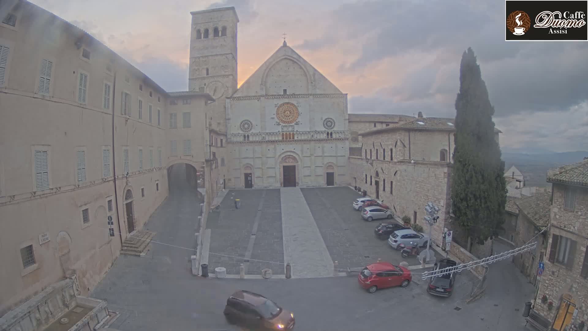 A large stone cathedral stands in a town square, under an overcast sky, with several cars parked nearby.