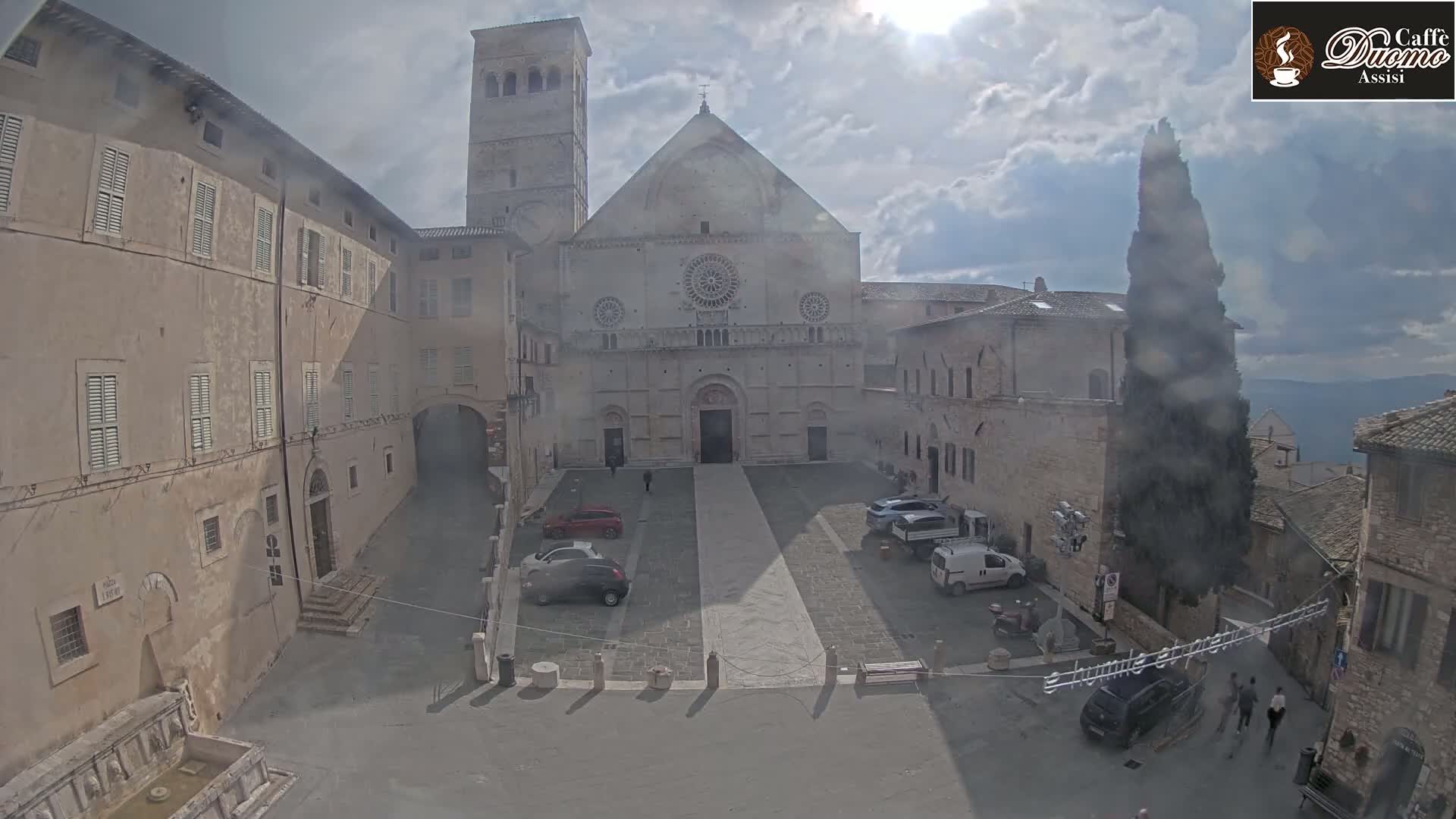 A sunny day reveals a large stone plaza with a cathedral at its center, surrounded by aged buildings, and several cars parked in the square.