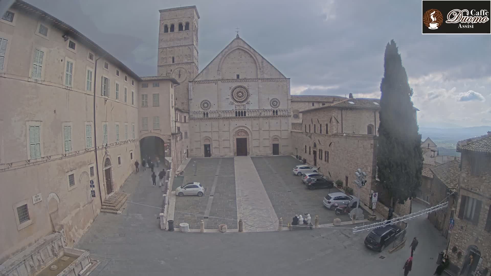 A large stone cathedral faces a paved plaza with several cars parked, surrounded by aged stone buildings under an overcast sky.