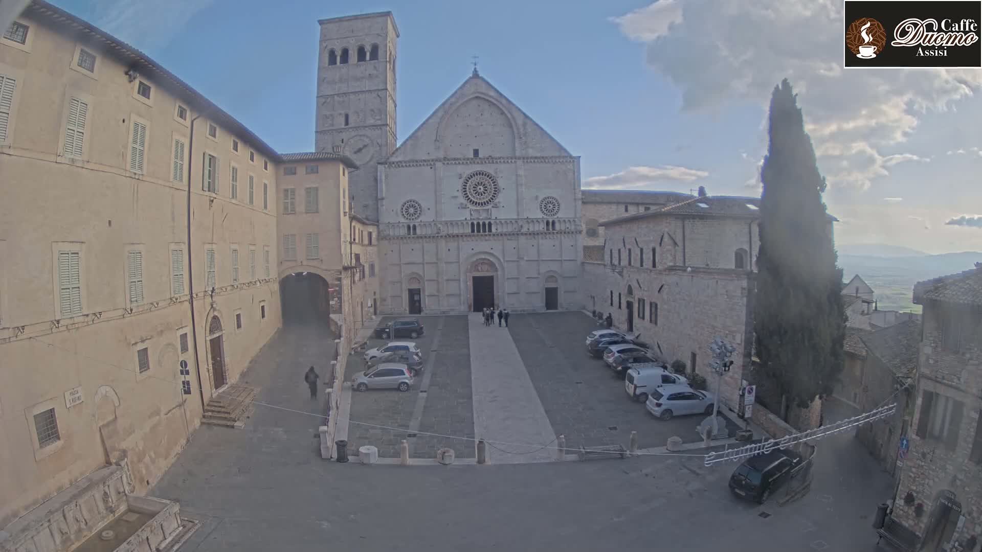 A large stone cathedral stands in a town square with several cars parked, under a partly cloudy sky.