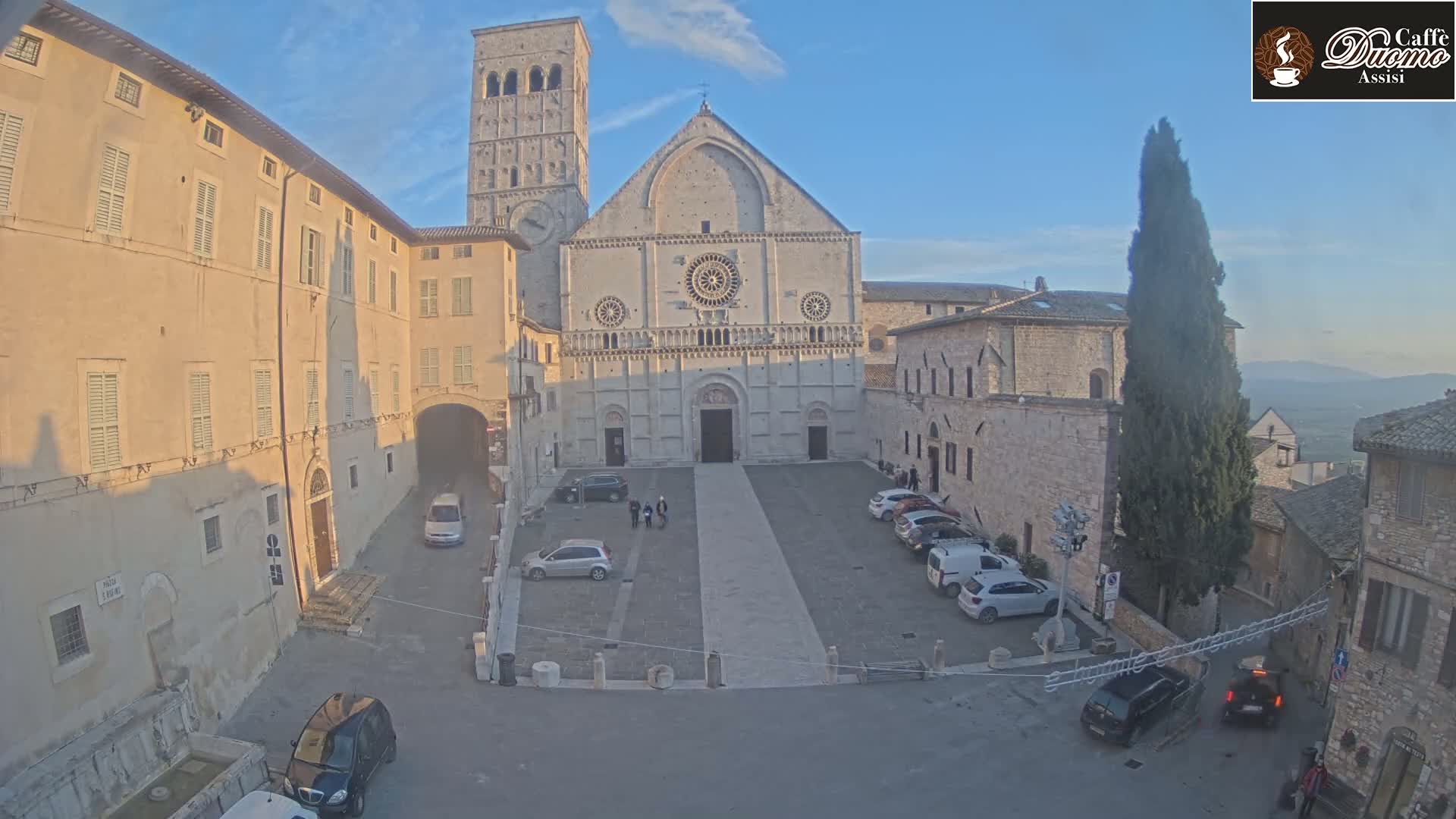 A large stone cathedral sits in a town square, surrounded by buildings and cars, under a clear, sunny sky.