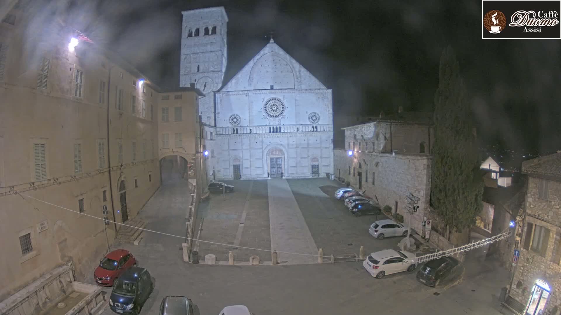 A nighttime view of a large stone cathedral in a town square, with several cars parked and a hazy atmosphere.
