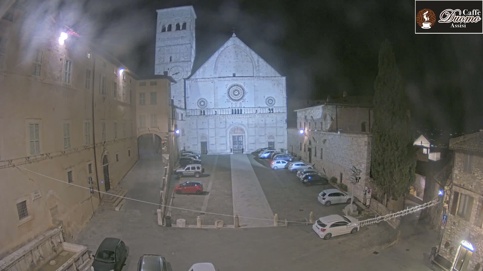 A large stone cathedral is at the center of a nighttime square, surrounded by buildings and parked cars under hazy conditions.