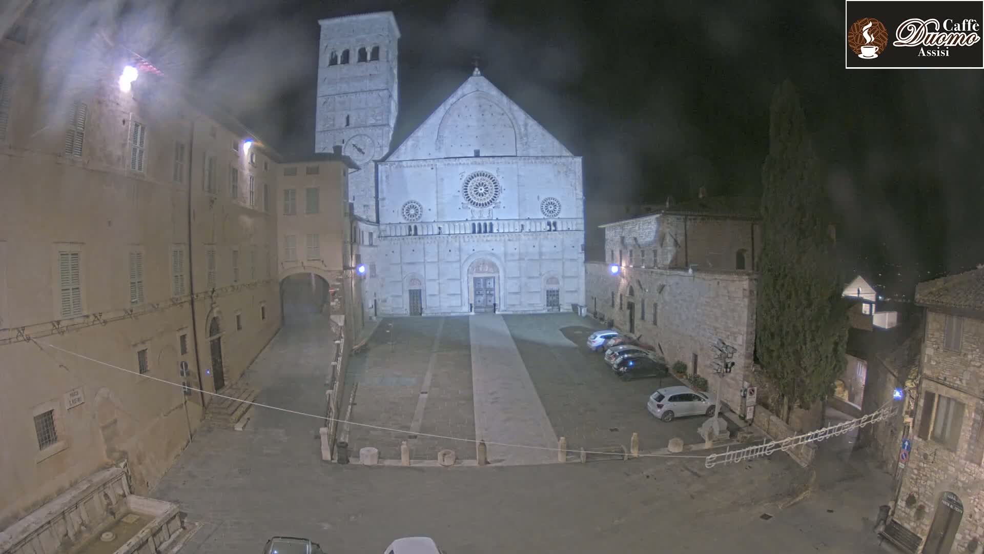 A nighttime view of a large, light-colored cathedral in a town square, with a few cars parked nearby under hazy conditions.