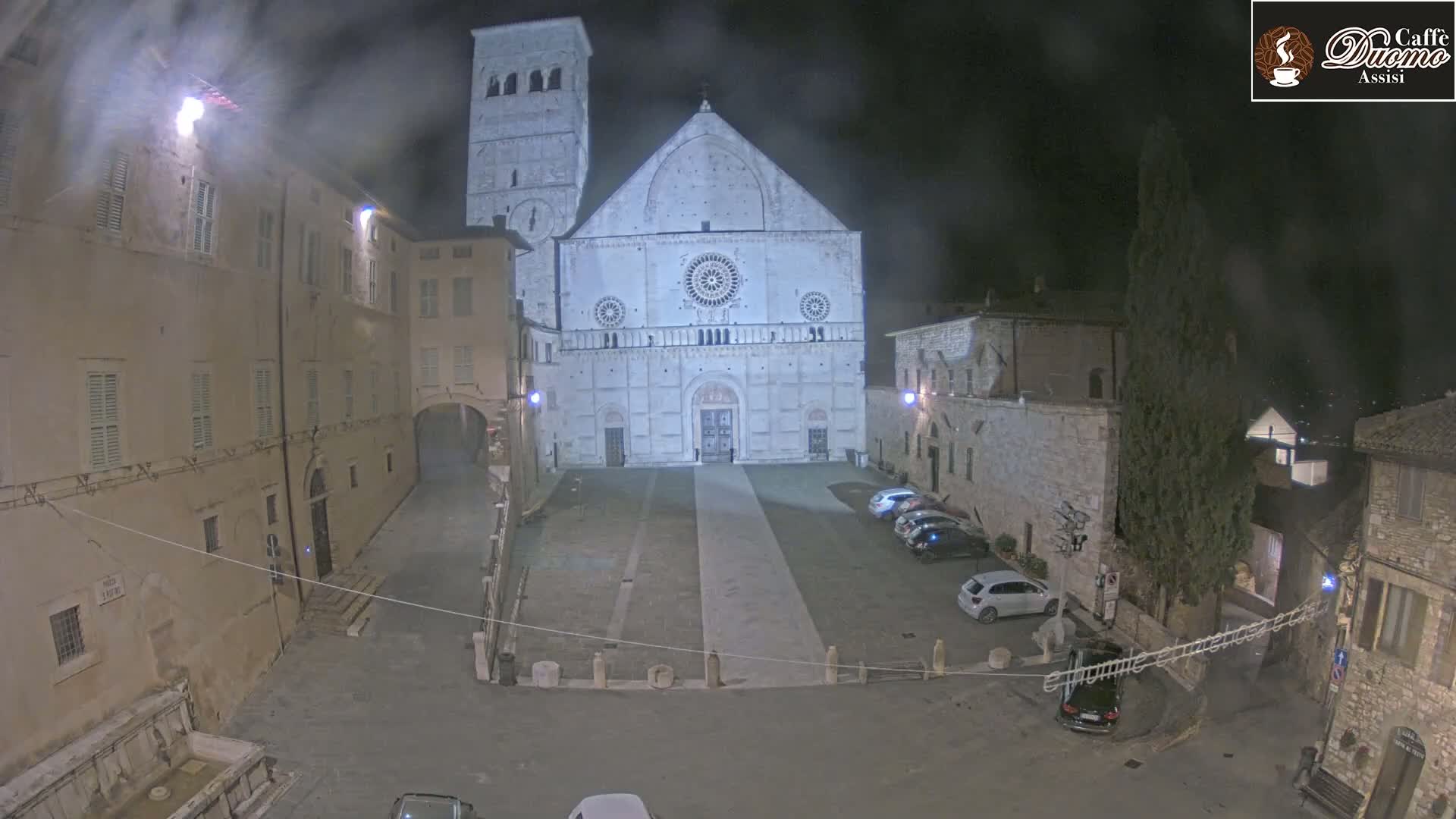A nighttime view of a stone plaza, with a large cathedral at its center, several parked cars, and surrounding buildings under hazy conditions.