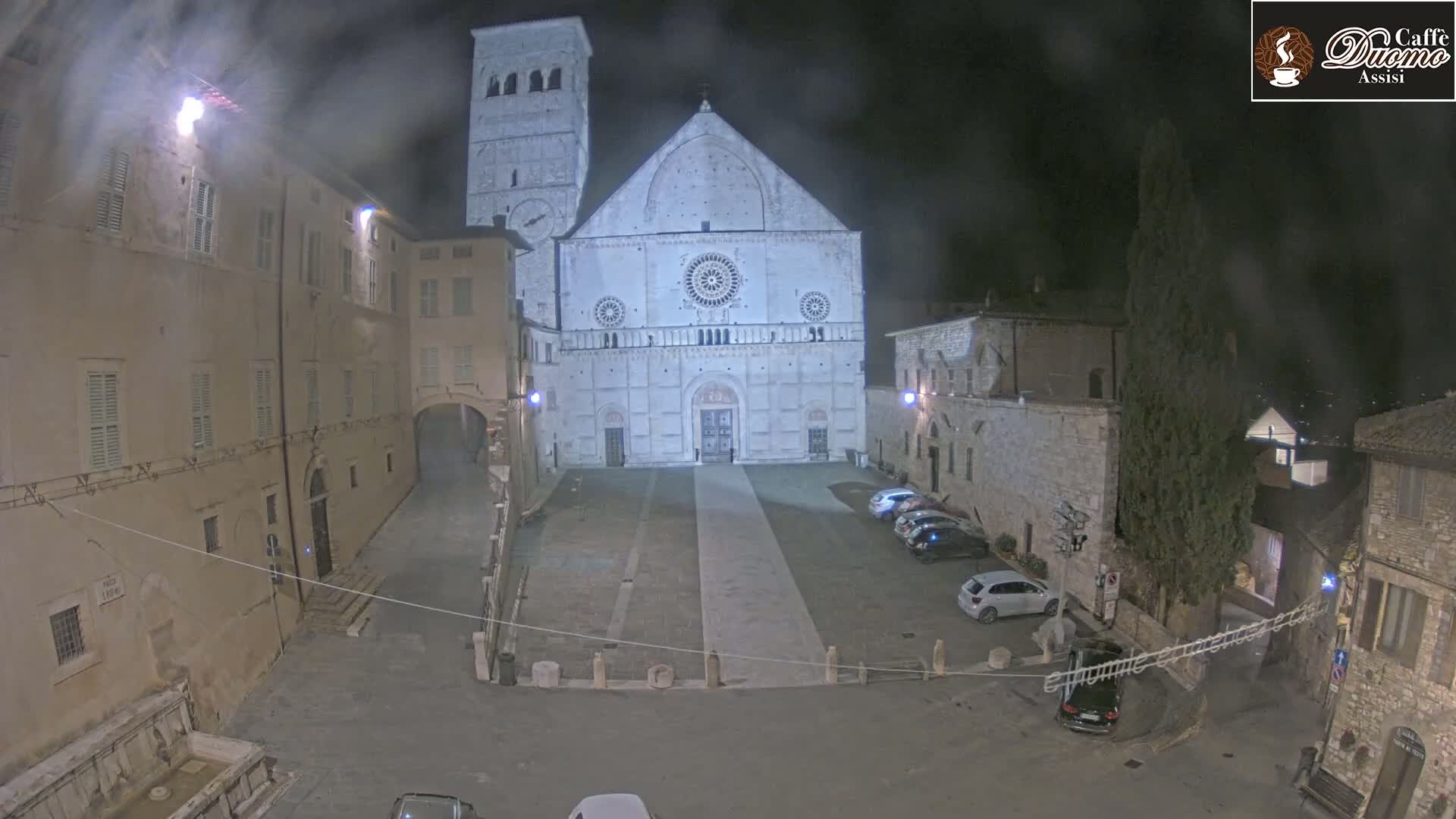 A large cathedral at night is surrounded by a town square with parked cars under hazy, dark conditions.