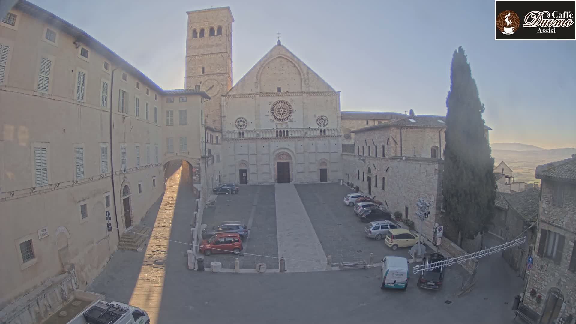A large stone cathedral with a bell tower stands in a town square with several cars parked, under a clear, bright sky.