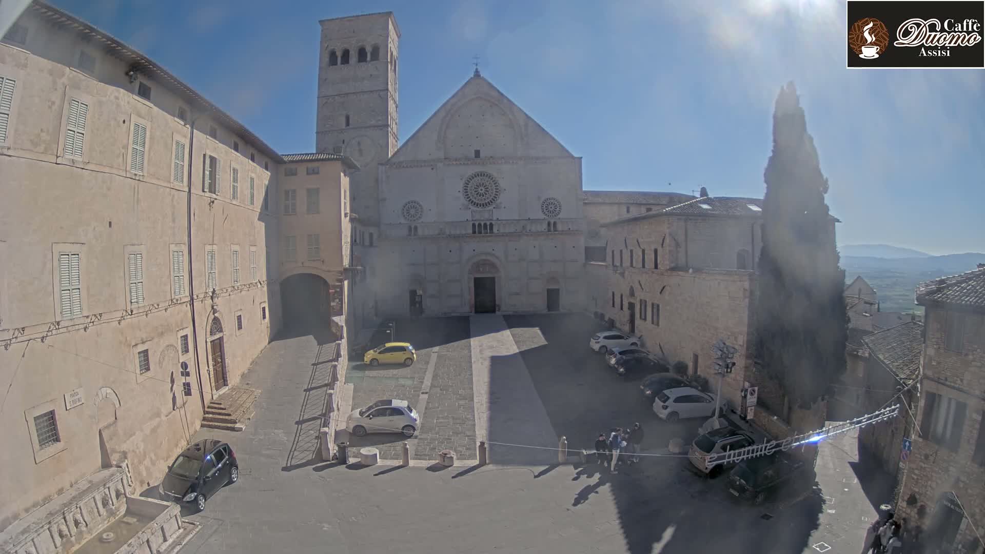A sunny, outdoor scene shows a large stone church and surrounding buildings in a town square with several parked cars and a few pedestrians.