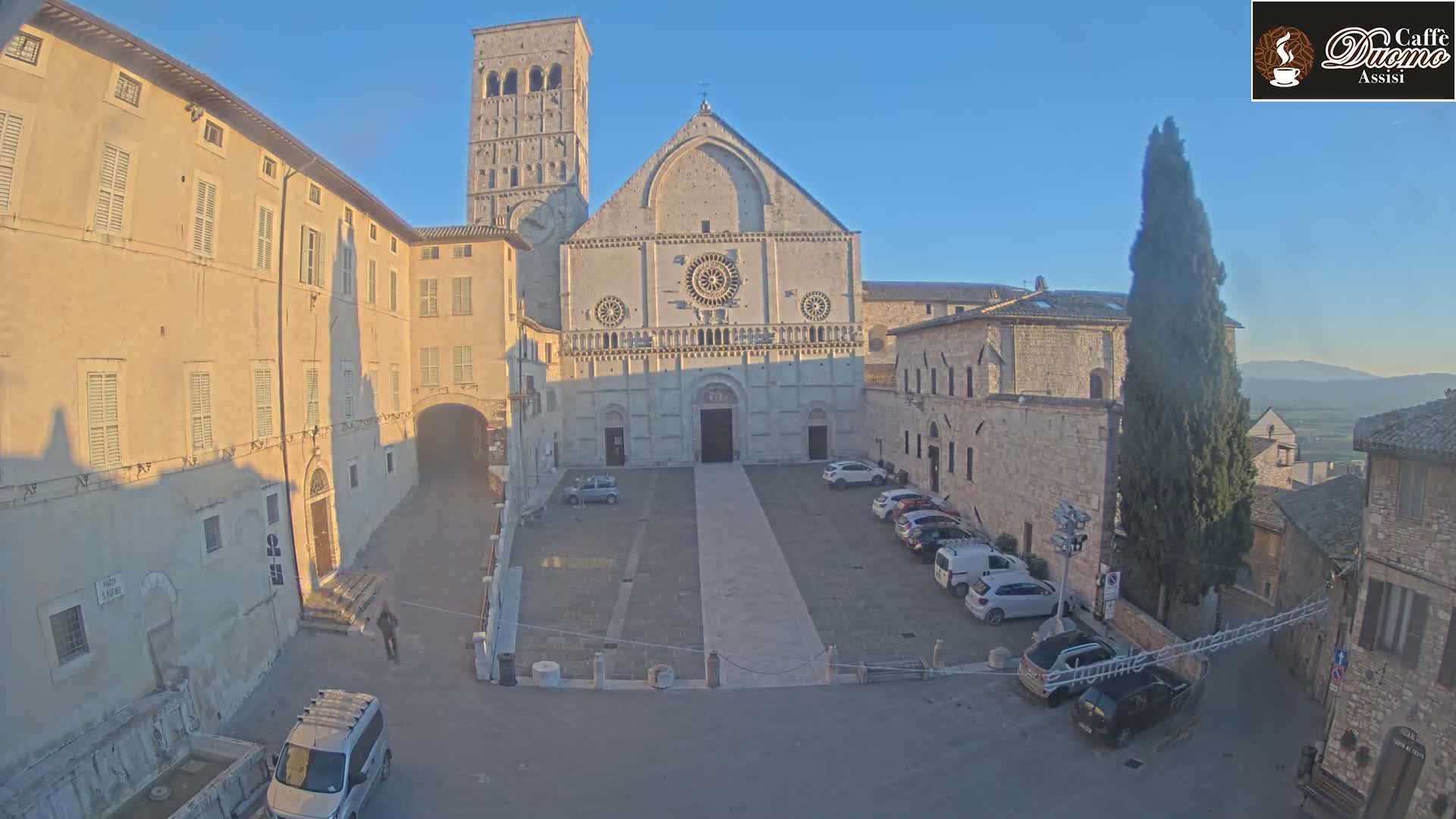 A large stone church with a bell tower stands in a town square with several cars parked, under a clear, bright sky.