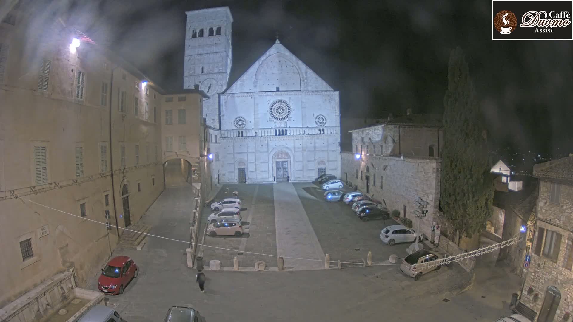 A large stone cathedral is visible at night in a town square with several cars parked, under a slightly hazy sky.