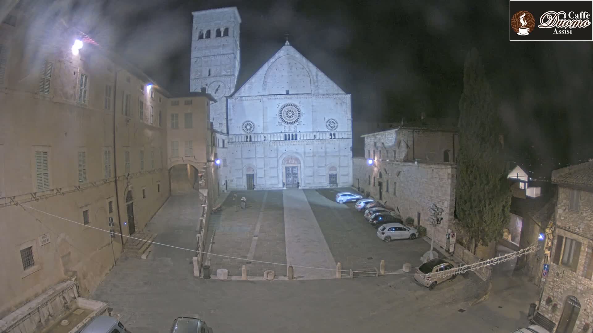 A large cathedral is illuminated at night in a town square with several parked cars and older buildings surrounding it under hazy conditions.