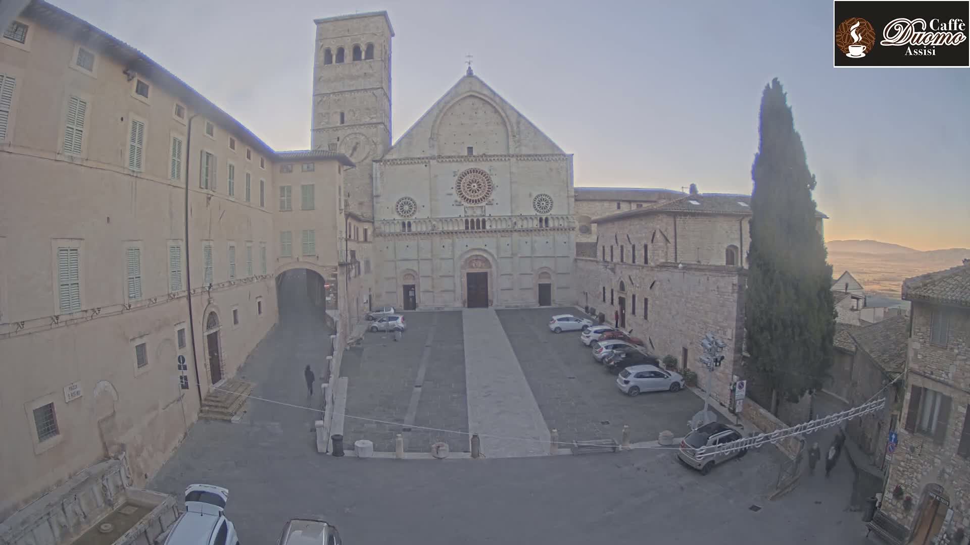 A large stone cathedral sits in a town square with several cars parked, under a twilight sky.