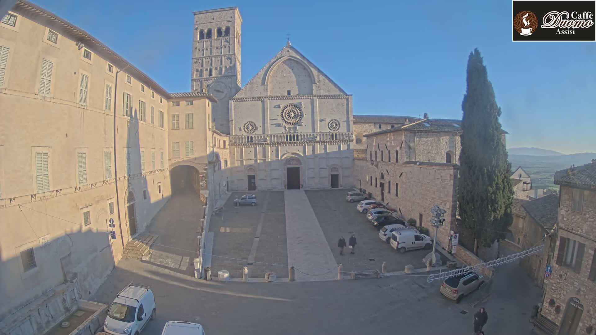 A large stone church with a bell tower stands in a sunny town square, surrounded by other stone buildings and a few parked cars.