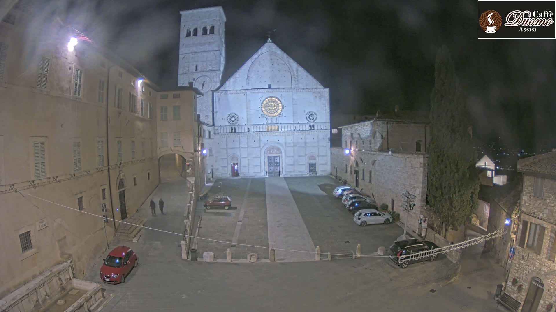 A large, light-colored cathedral stands in a mostly empty, stone plaza at night, under a hazy sky, with several cars parked around the perimeter.