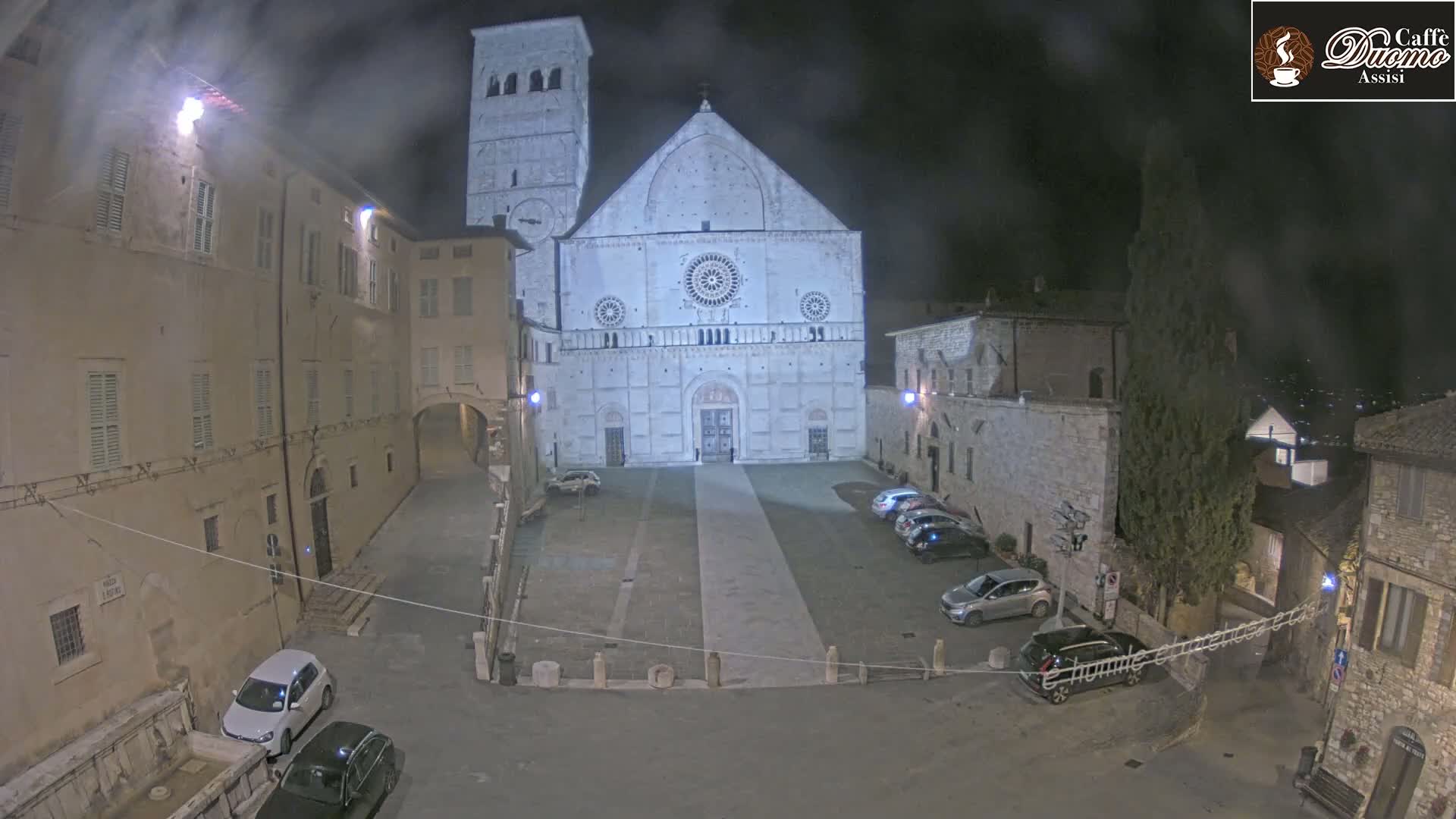 A large cathedral, illuminated at night, sits in a town square with several cars parked and older buildings surrounding it under hazy or foggy conditions.