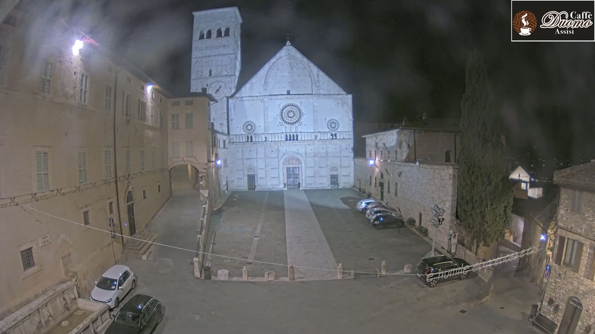 A nighttime view of a large cathedral in a town square, with a few cars parked and a misty or hazy atmosphere.