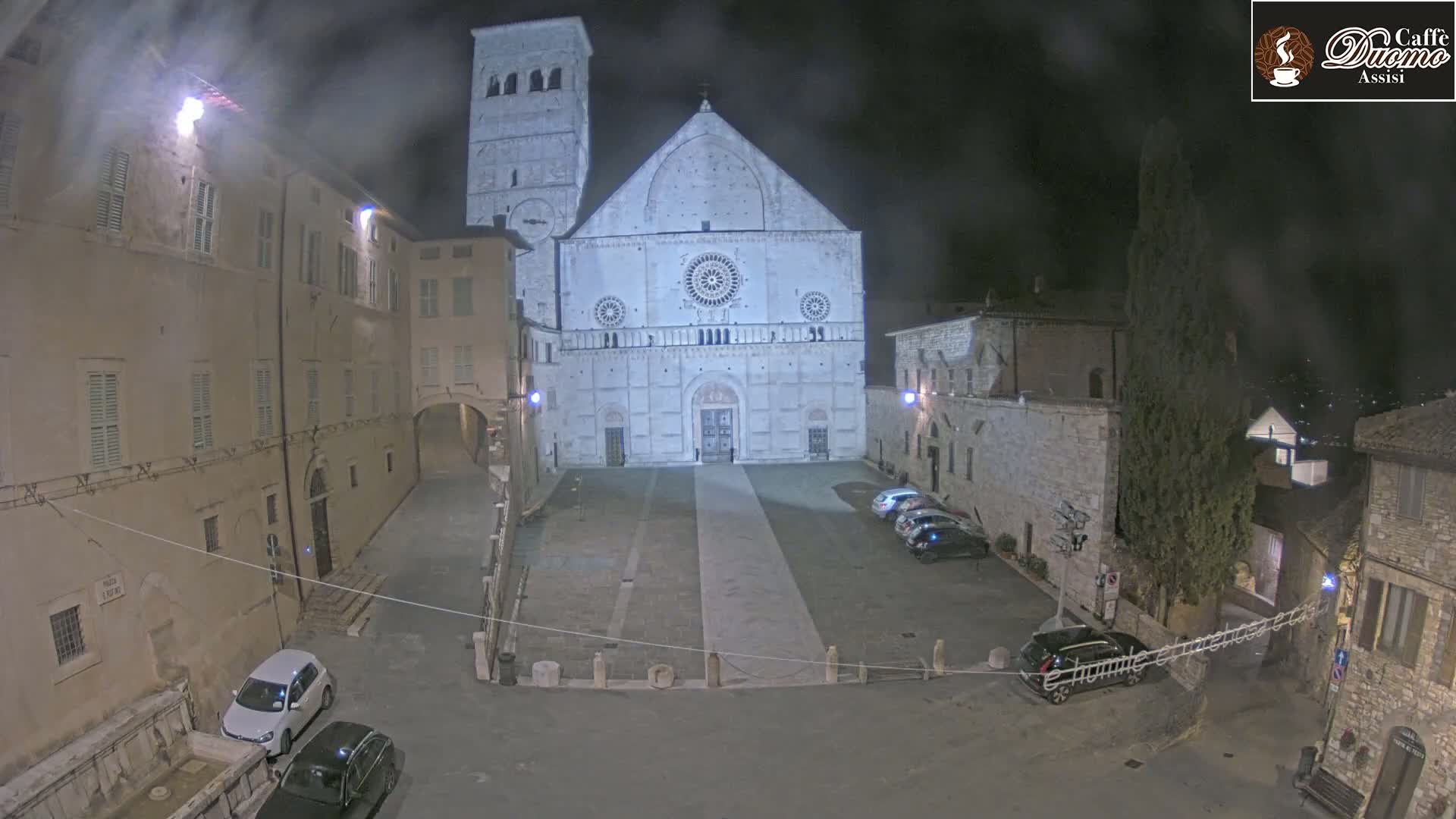 A nighttime view of a town square with a large stone cathedral at its center, under a hazy sky.